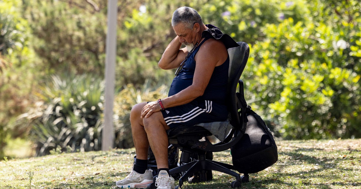 A man sitting in a black office chair in a shady park wipes his neck. He wears shorts, sneakers and a tank top.