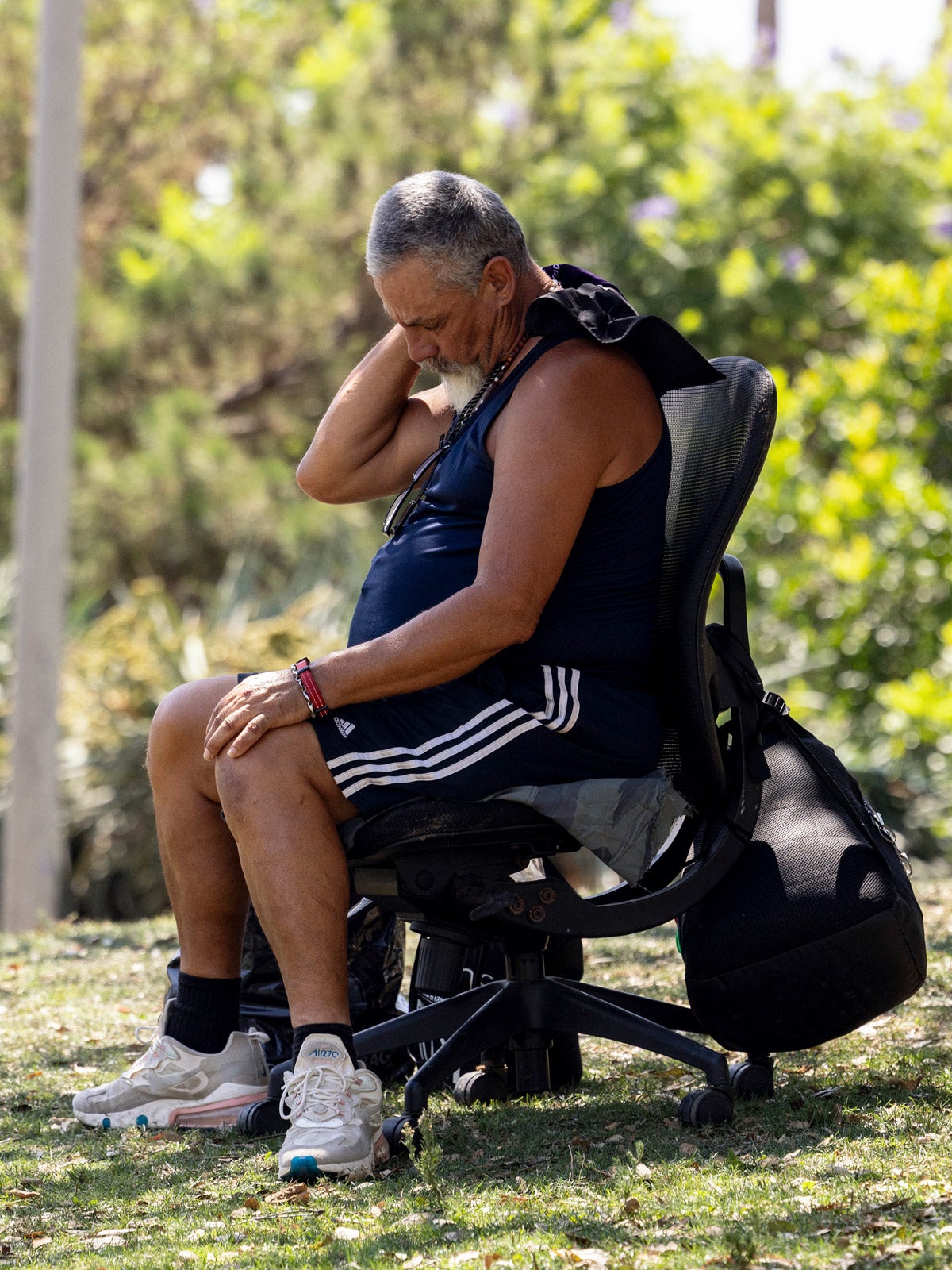 A man sitting in a black office chair in a shady park wipes his neck. He wears shorts, sneakers and a tank top.