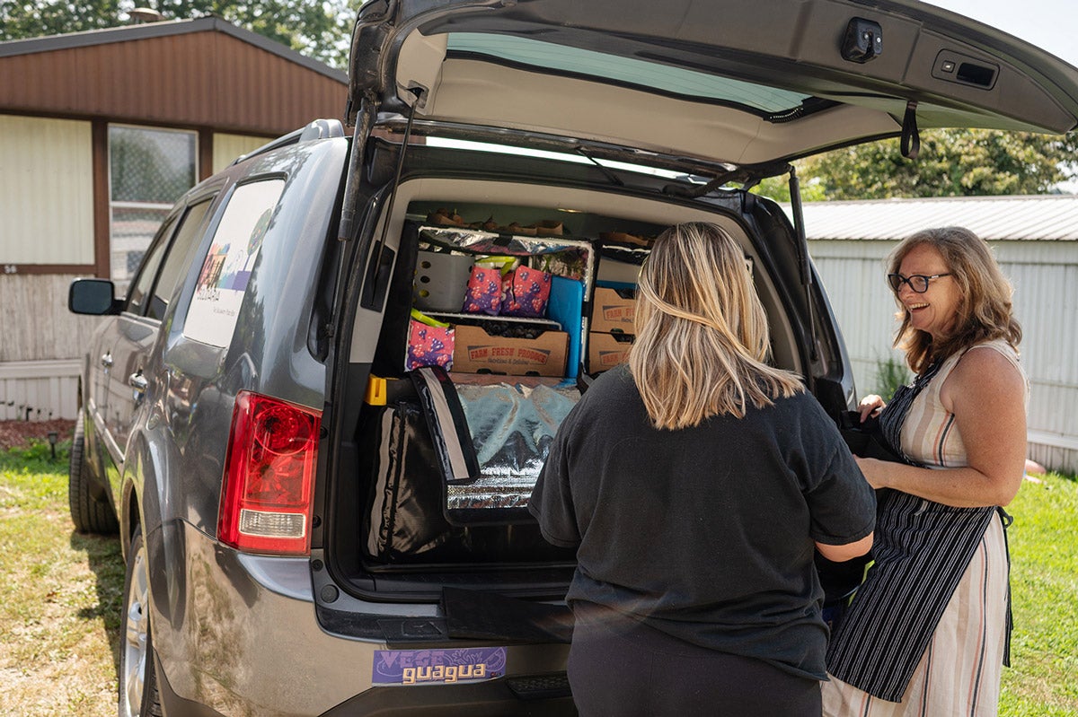 Two women chat behind an open black SUV trunk loaded with food boxes.