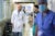 A bald, male physician and a male PA walk down a hospital hallway. A surgeon in scrubs and a mask crossed in the foreground.