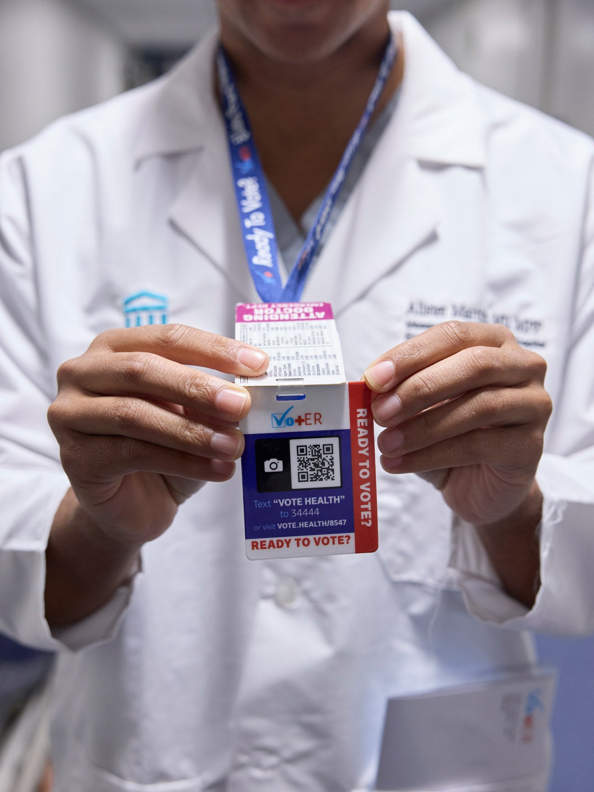 A physician holds a "VOT-ER" card in his hand that's attached to a lanyard. The card has a QR code, text instructions and the words "Ready to Vote?"