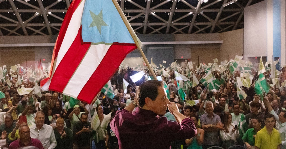 Politician Juan Dalmau, photographed from behind, waves a Puerto Rico flag in front of a large crowd of supporters at a convention.