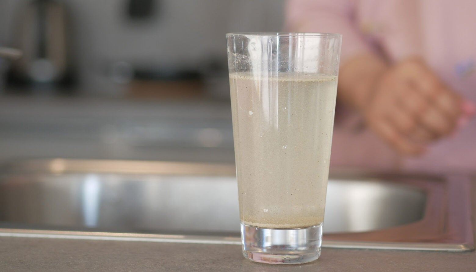 A glass of dirty water stands on a kitchen counter near a sink. A blurred person in a pink shirt is in the background