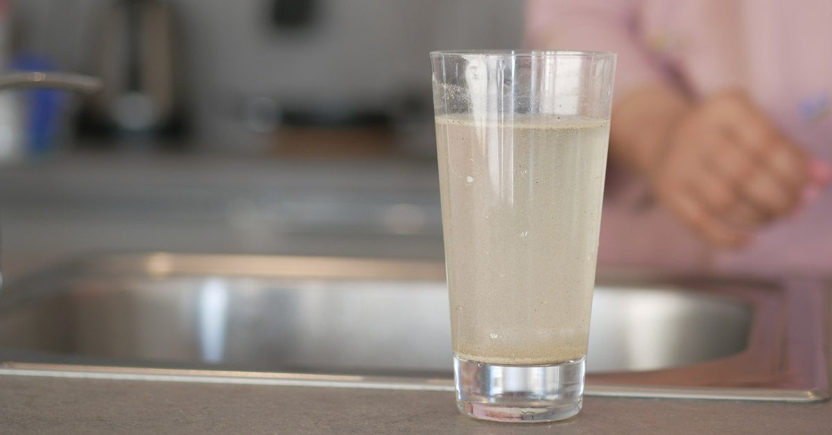 A glass of dirty water stands on a kitchen counter near a sink. A blurred person in a pink shirt is in the background