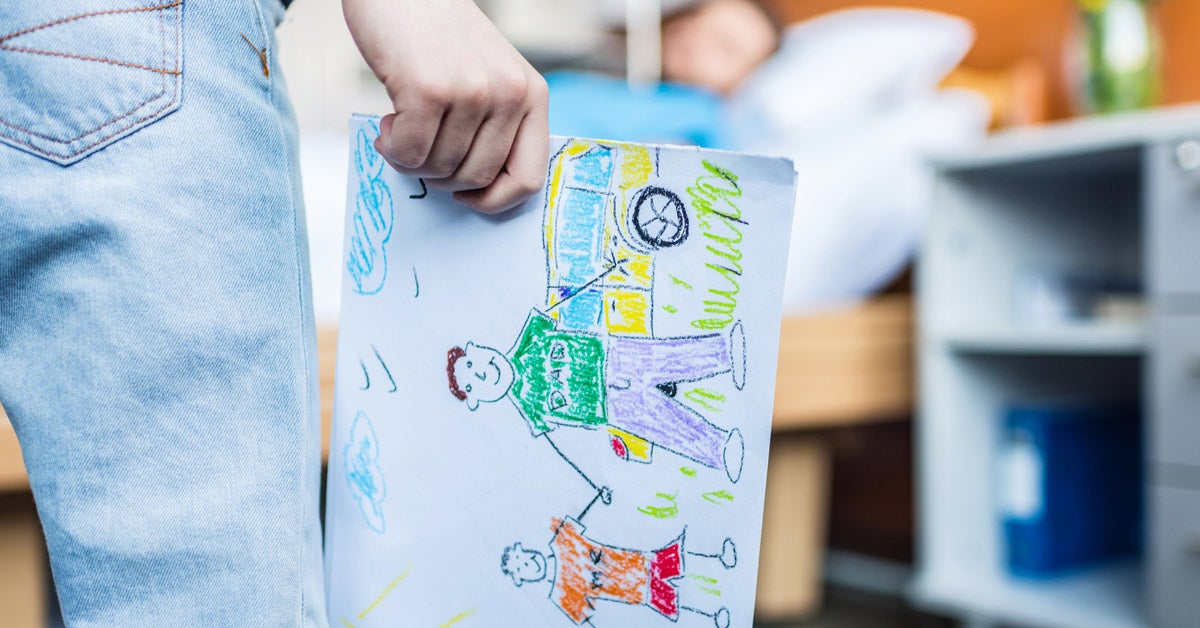 A child holds a self-made drawing in one hand while I parent rests in a facility bed in the blurred background. The illustration is of the child and parent holding hands on a sunny day by a yellow car.