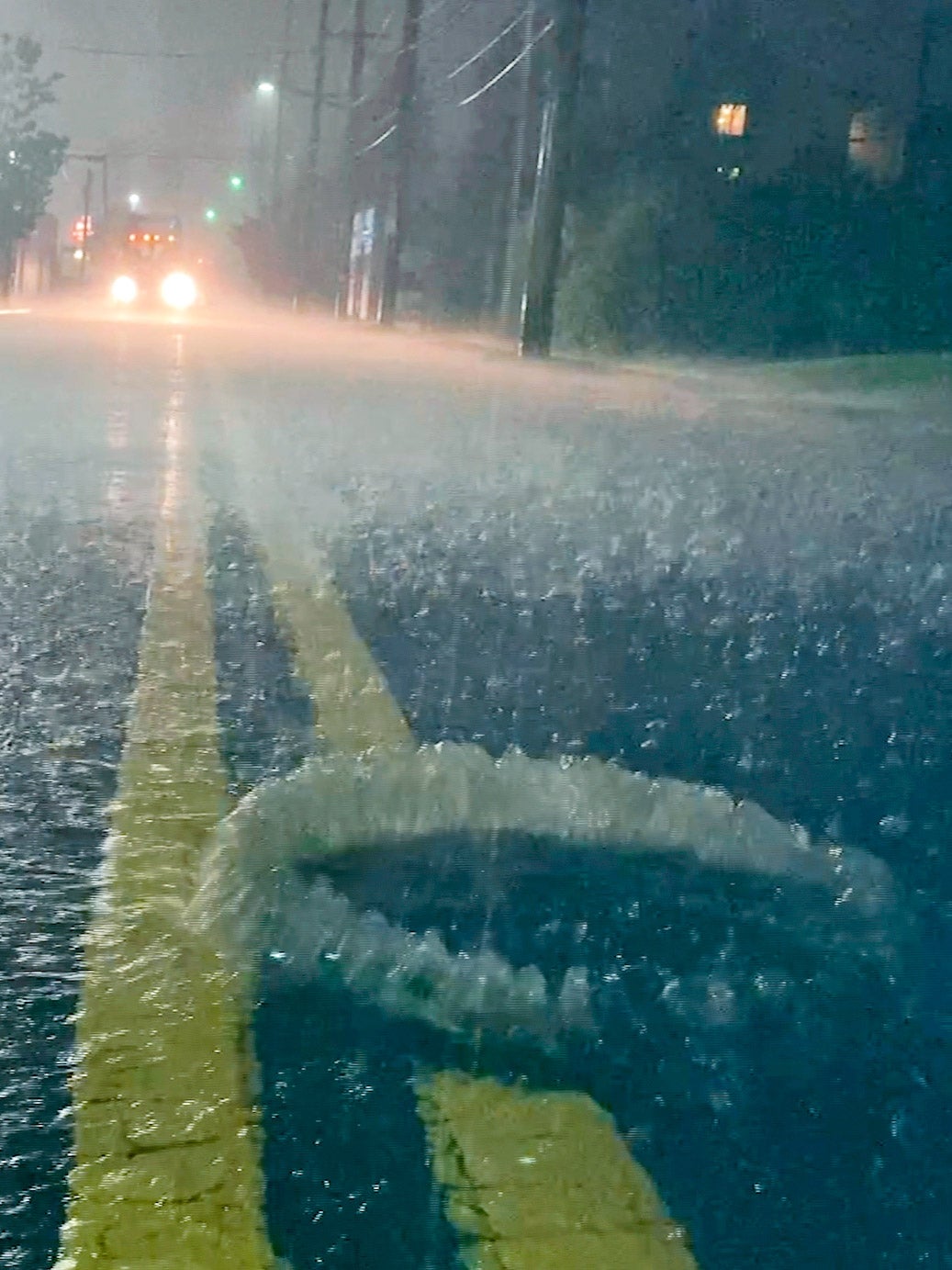 A flooded neighborhood street with a utility hole overflowing with bubbles. Car with headlights approaches.