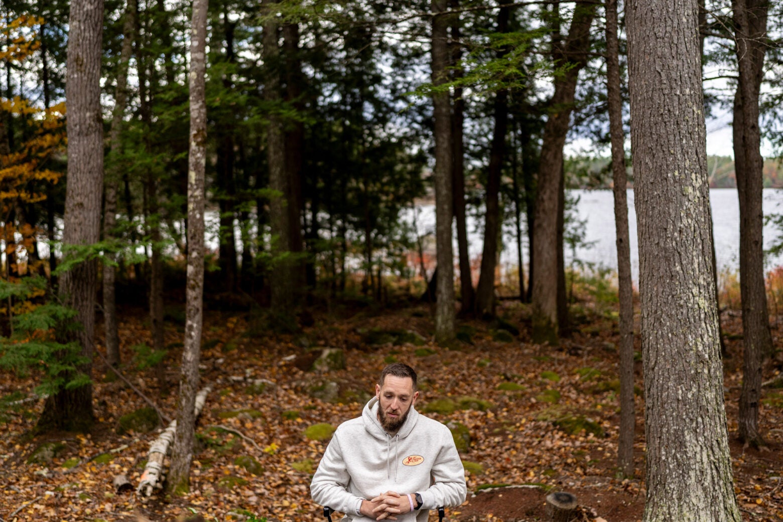 A middle-aged man wearing a hoodie sits in a chair near a pine forest and lake. He has his hands clasped in his lap and looks down.