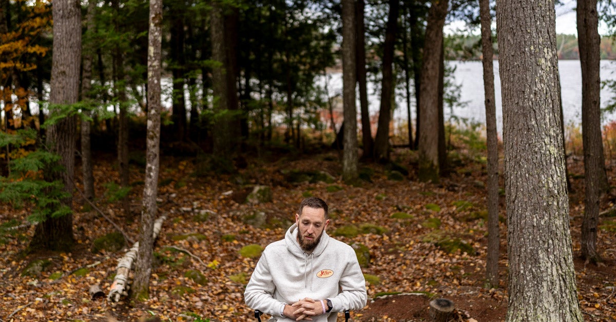 A middle-aged man wearing a hoodie sits in a chair near a pine forest and lake. He has his hands clasped in his lap and looks down.