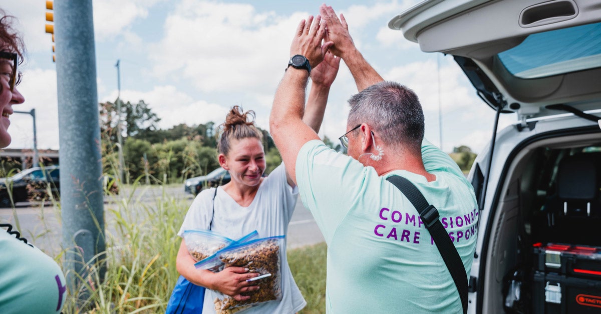 A man and a woman high-five along a highway in Durham, NC. One person holds zip-lock bags of dog food and her phone. The other wears a t-shirt that says "compassion care response."