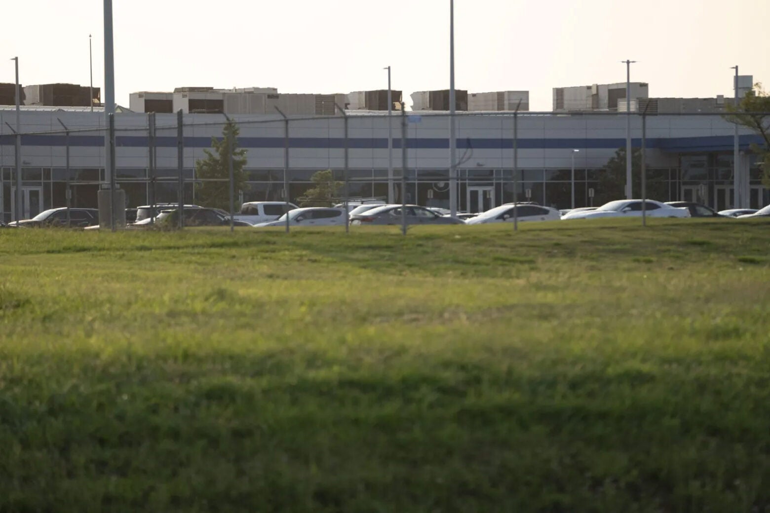 Cars parked alongside a facility in Memphis. There are multiple grey cooling rowers on the roof. A green field and fence are in the foreground of the image.