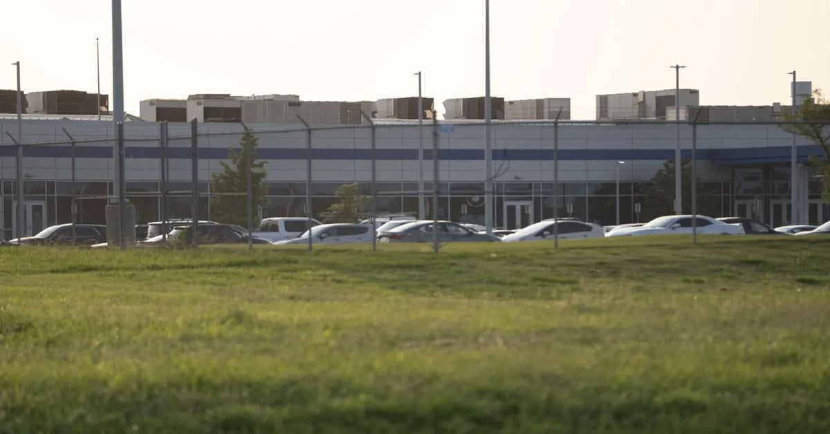 Cars parked alongside a facility in Memphis. There are multiple grey cooling rowers on the roof. A green field and fence are in the foreground of the image.