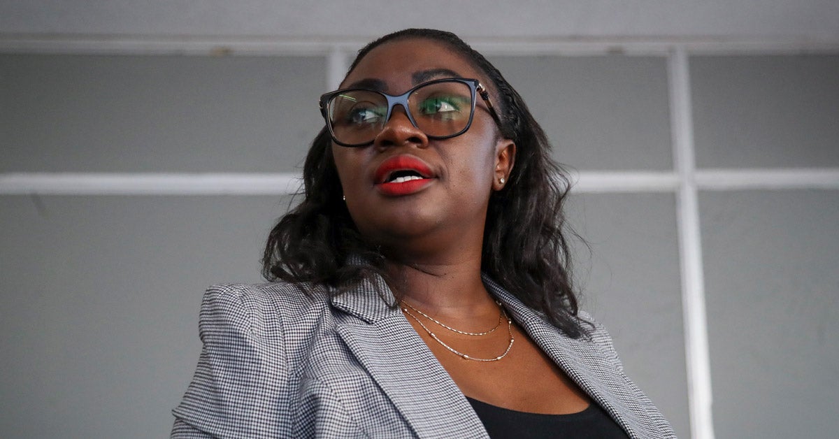 Kenyan senator Gloria Orwoba speaks at a press conference. She wears a grey striped blazer, glasses and red lipstick.