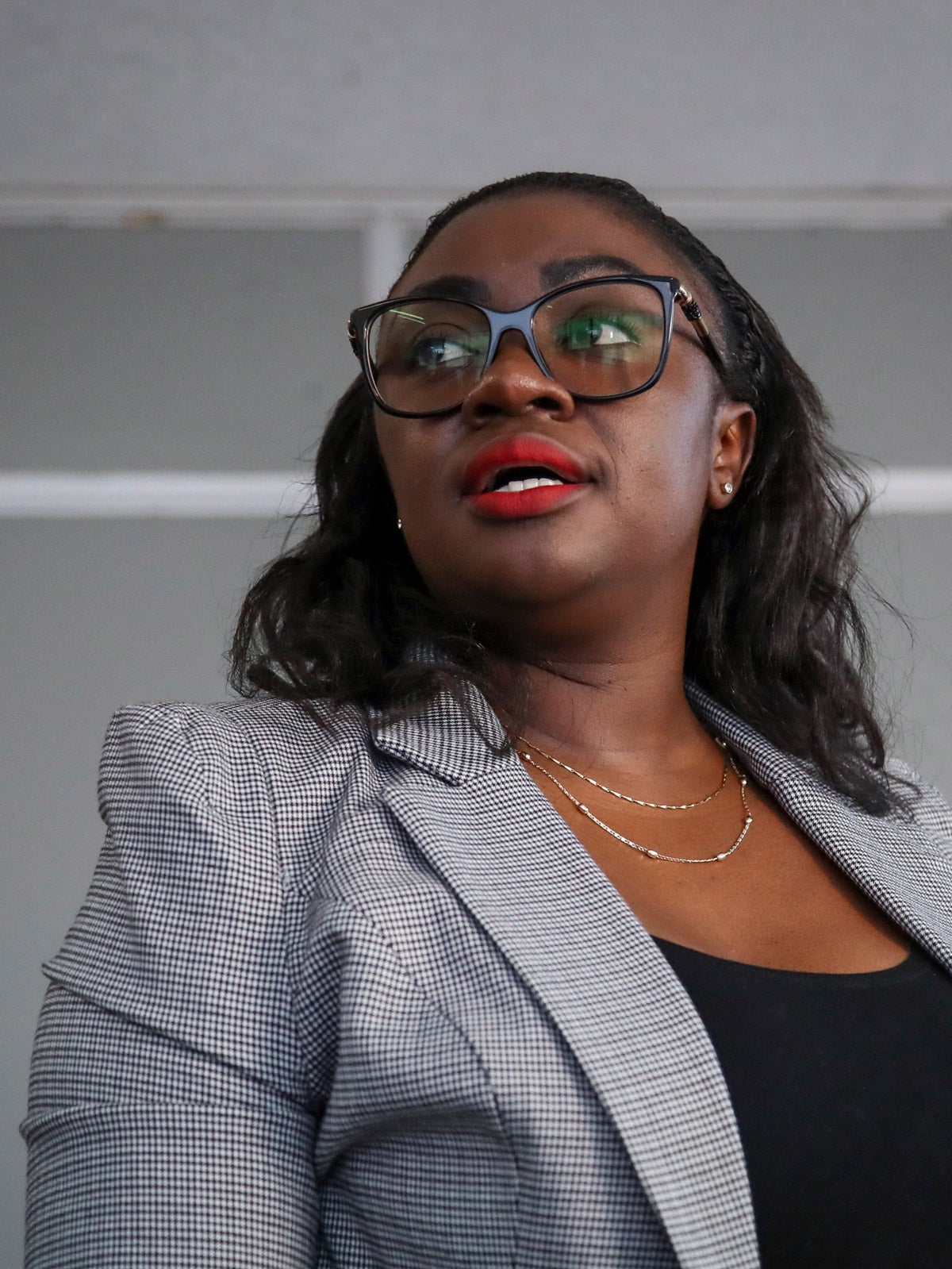 Kenyan senator Gloria Orwoba speaks at a press conference. She wears a grey striped blazer, glasses and red lipstick.