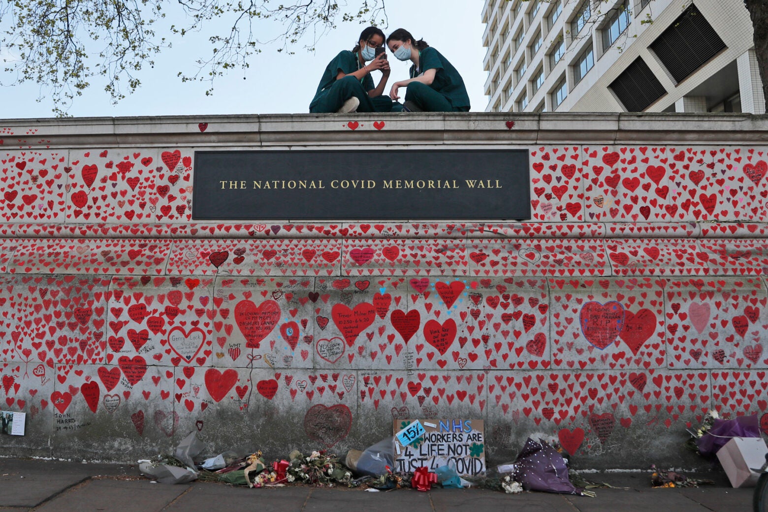 Two nurses in dark teal scrubs and masks sit on top of The National COVID Memorial Wall in London. The wall is covered with handrawn red hearts and messages.