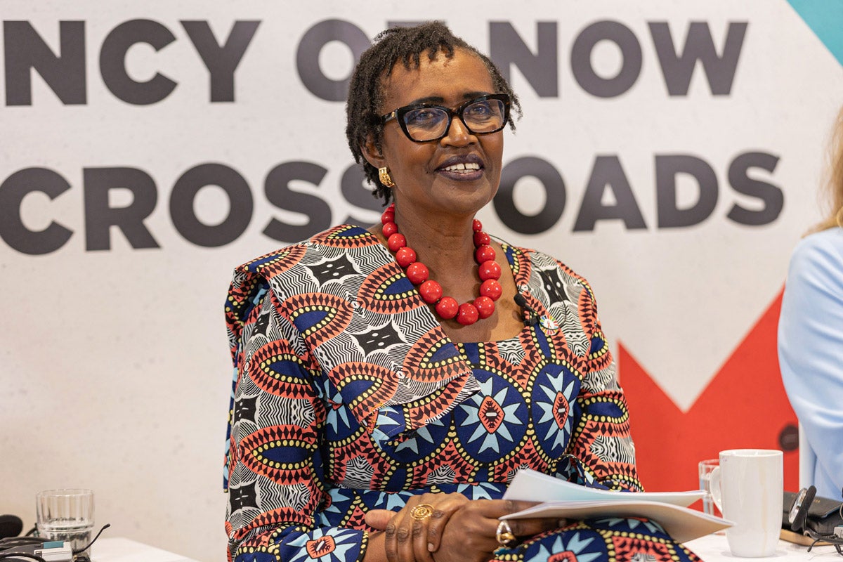 Winnie Byanyima sits on a panel at a conference. She holds papers in her lap and smiles at the audience.