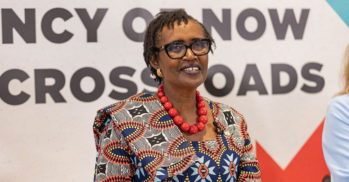 Winnie Byanyima sits on a panel at a conference. She holds papers in her lap and smiles at the audience.