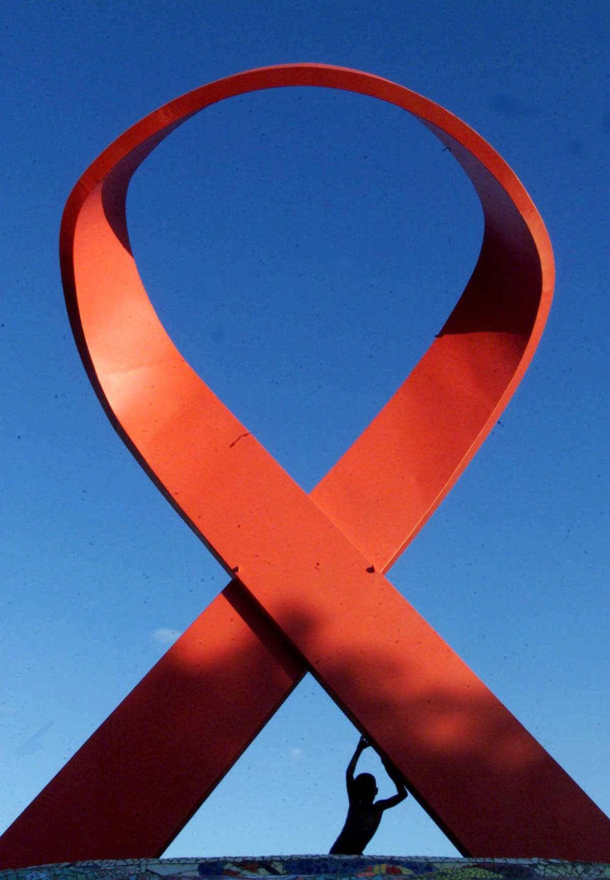 A giant AIDS red ribbon statue. A young silhouetted child pushes against one strand.